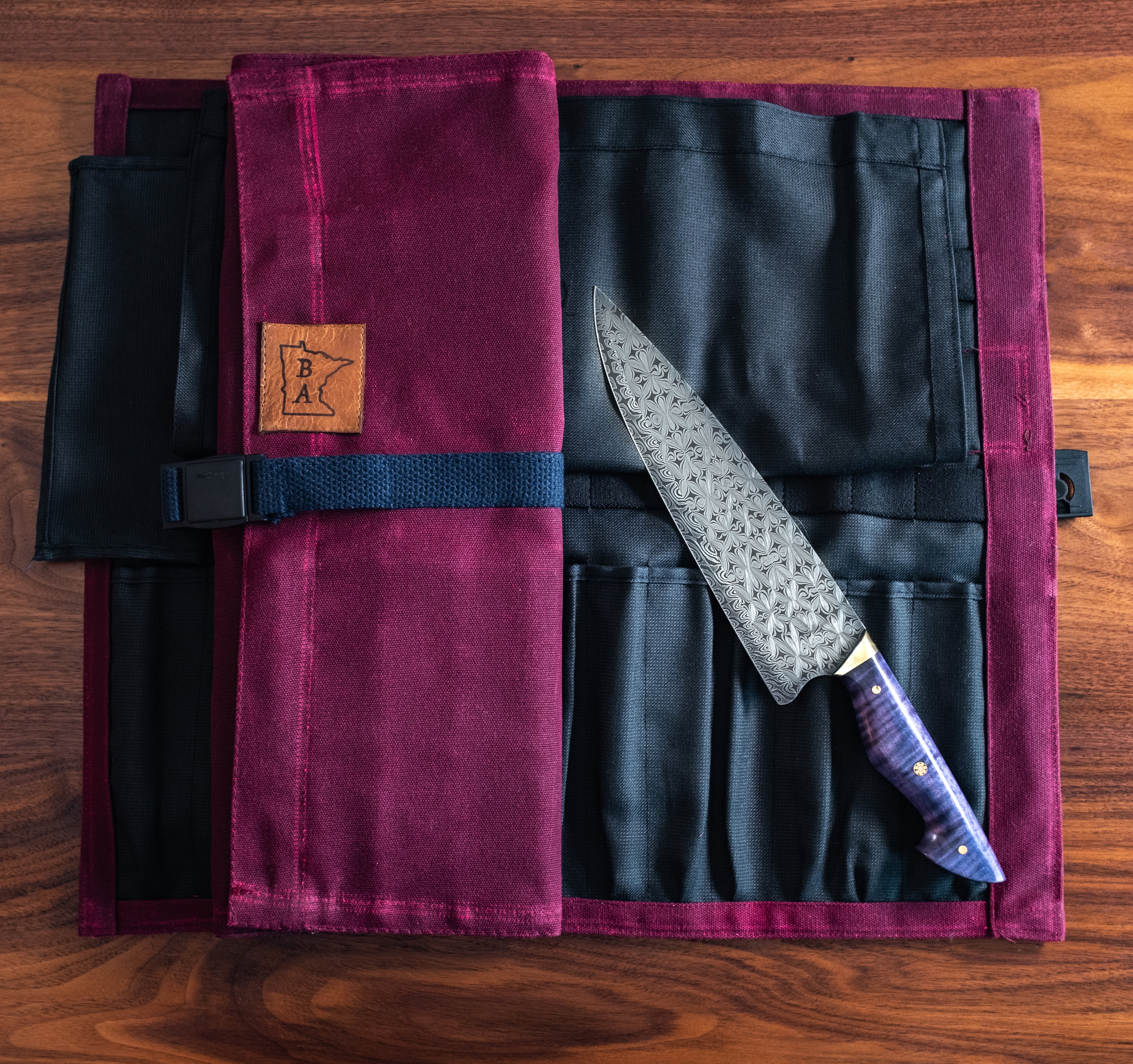 Burgundy Side Hustle knife roll unfolded showing the kevlar interior. Another burgundy Side Hustle knife roll sitting on top alongside a knife. Produced in Minnesota by Craftmade Aprons.