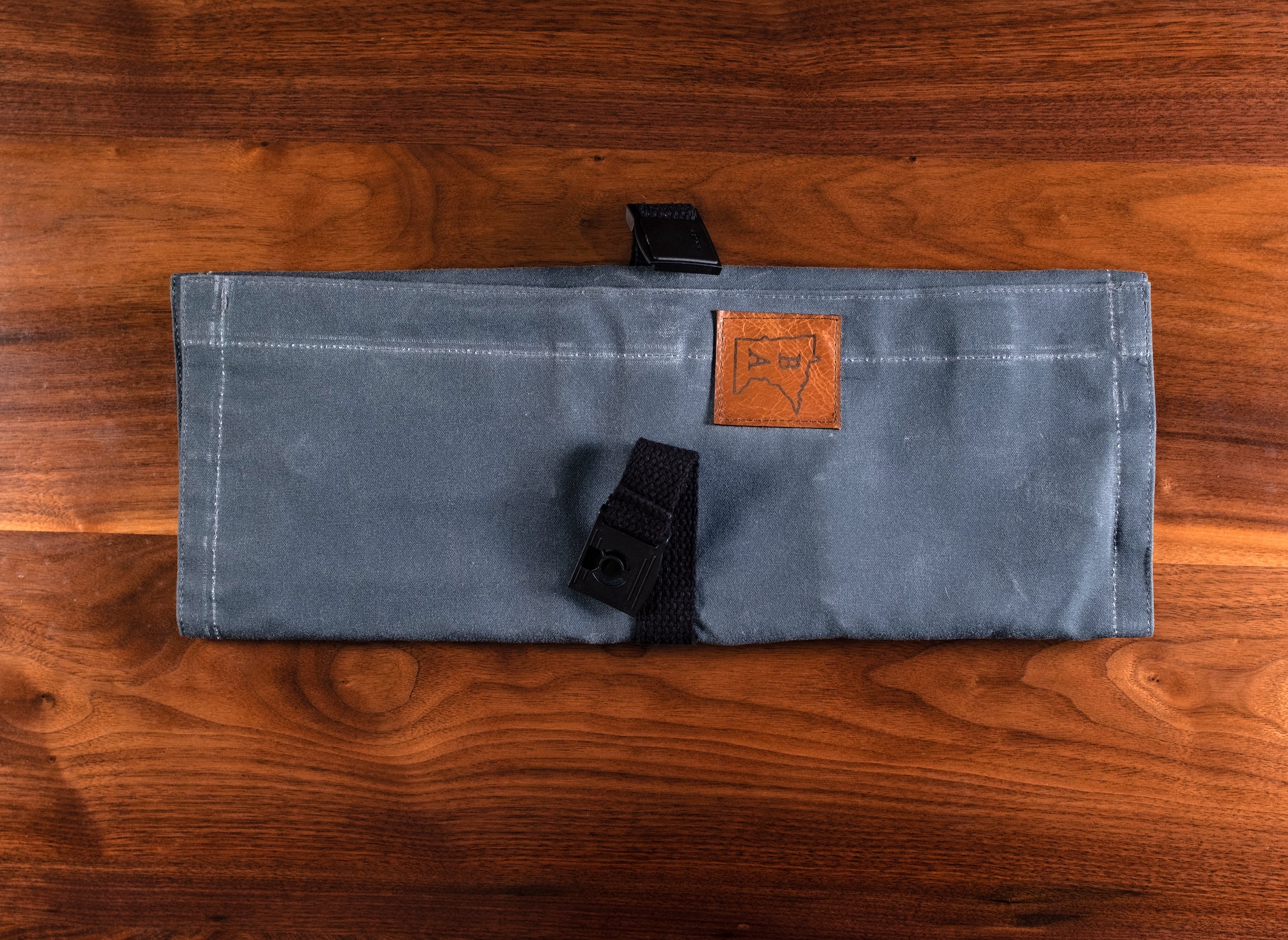 The slate Side Hustle knife roll, designed by Craftmade Aprons in Minnesota, laying unbuckled on a wooden surface.