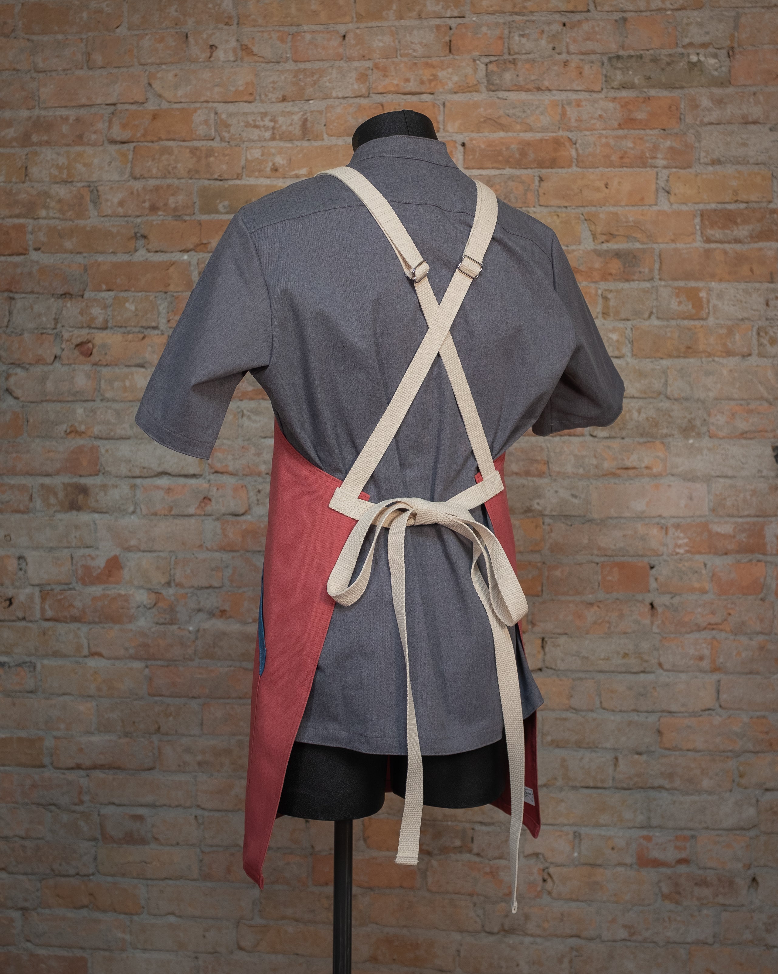 Cotton duck cloth crossback apron from Craftmade Aprons, Minnesota, displayed on a mannequin over a gray chef coat. The apron design Sockeye is shown from the back, displaying the tan crossback strapping.