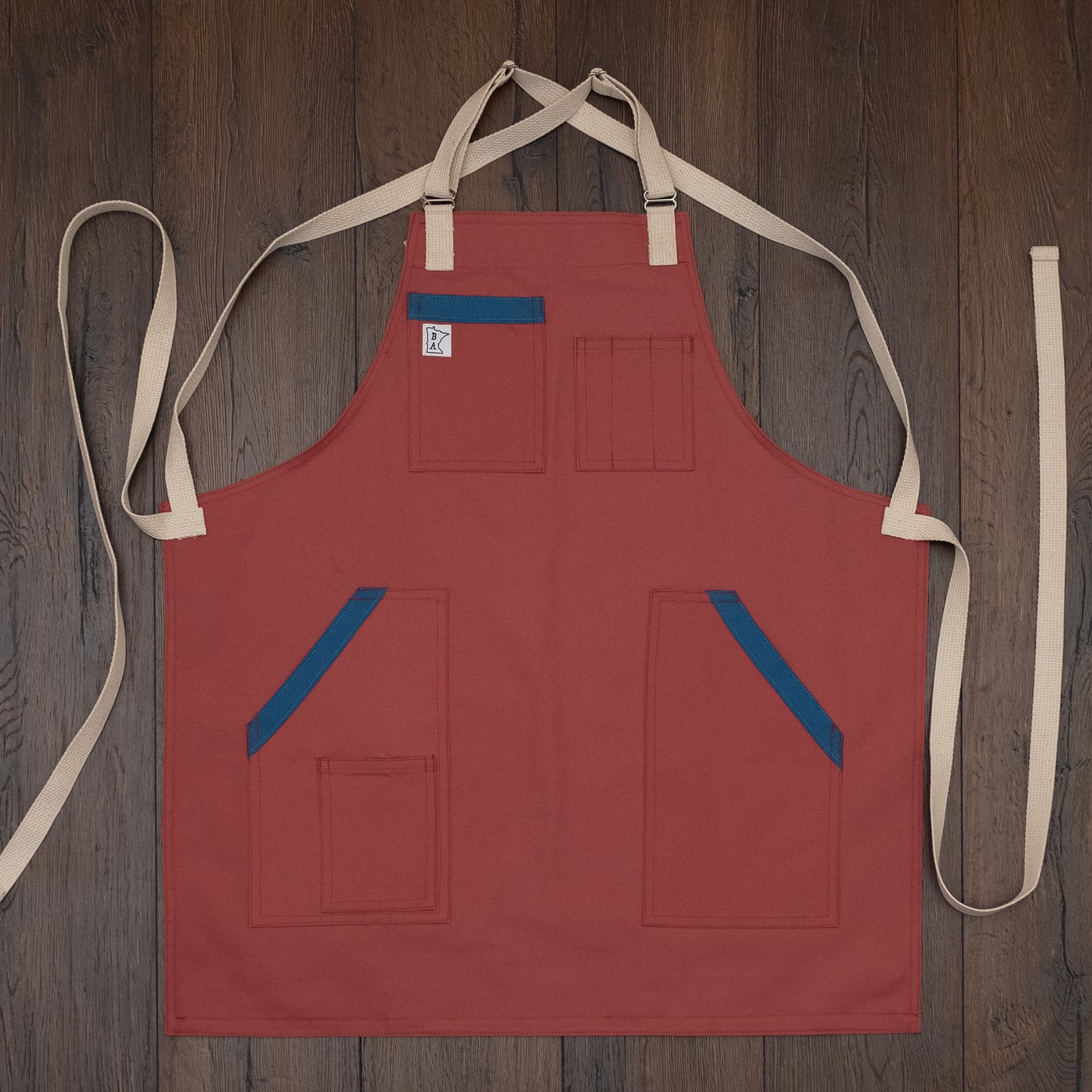 Pink-salmon colored duck cloth apron with blue accents on the pockets laid out flat on a wooden surface. Design Sockeye by Craftmade Aprons, based in Minnesota.