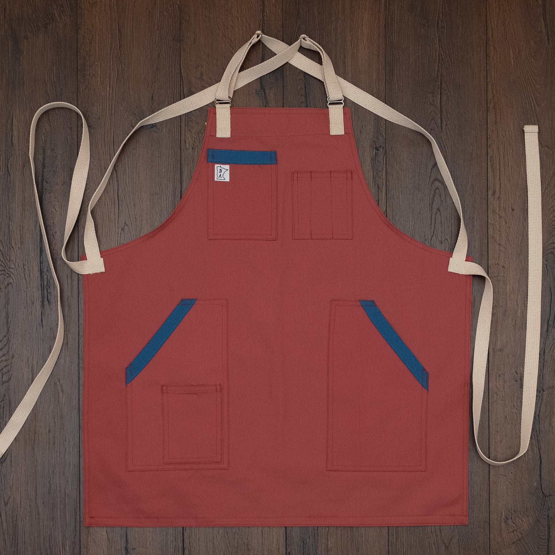 Pink-salmon colored duck cloth apron with blue accents on the pockets laid out flat on a wooden surface. Design Sockeye by Craftmade Aprons, based in Minnesota.