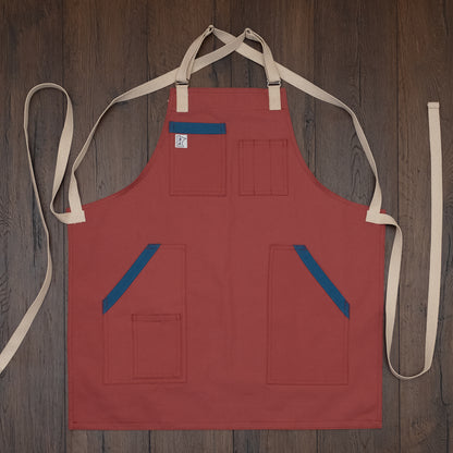 Pink-salmon colored duck cloth apron with blue accents on the pockets laid out flat on a wooden surface. Design Sockeye by Craftmade Aprons, based in Minnesota.