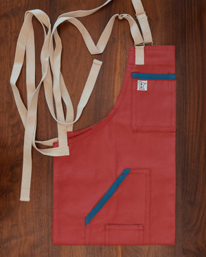 Pink-salmon cotton duck cloth apron with blue trim and tan strapping folded on a wooden surface. The apron design Sockeye was manufactured in Minnesota by Craftmade Aprons.