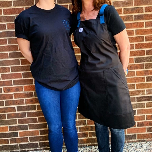 Two individuals wearing products from the Black and Blue Collection. Black and Blue T-shirt worn by the model on the left and Black and Blue Apron worn by the model on the right. Both the T-shirt and the apron were manufactured on Minnesota by Craftmade Aprons.