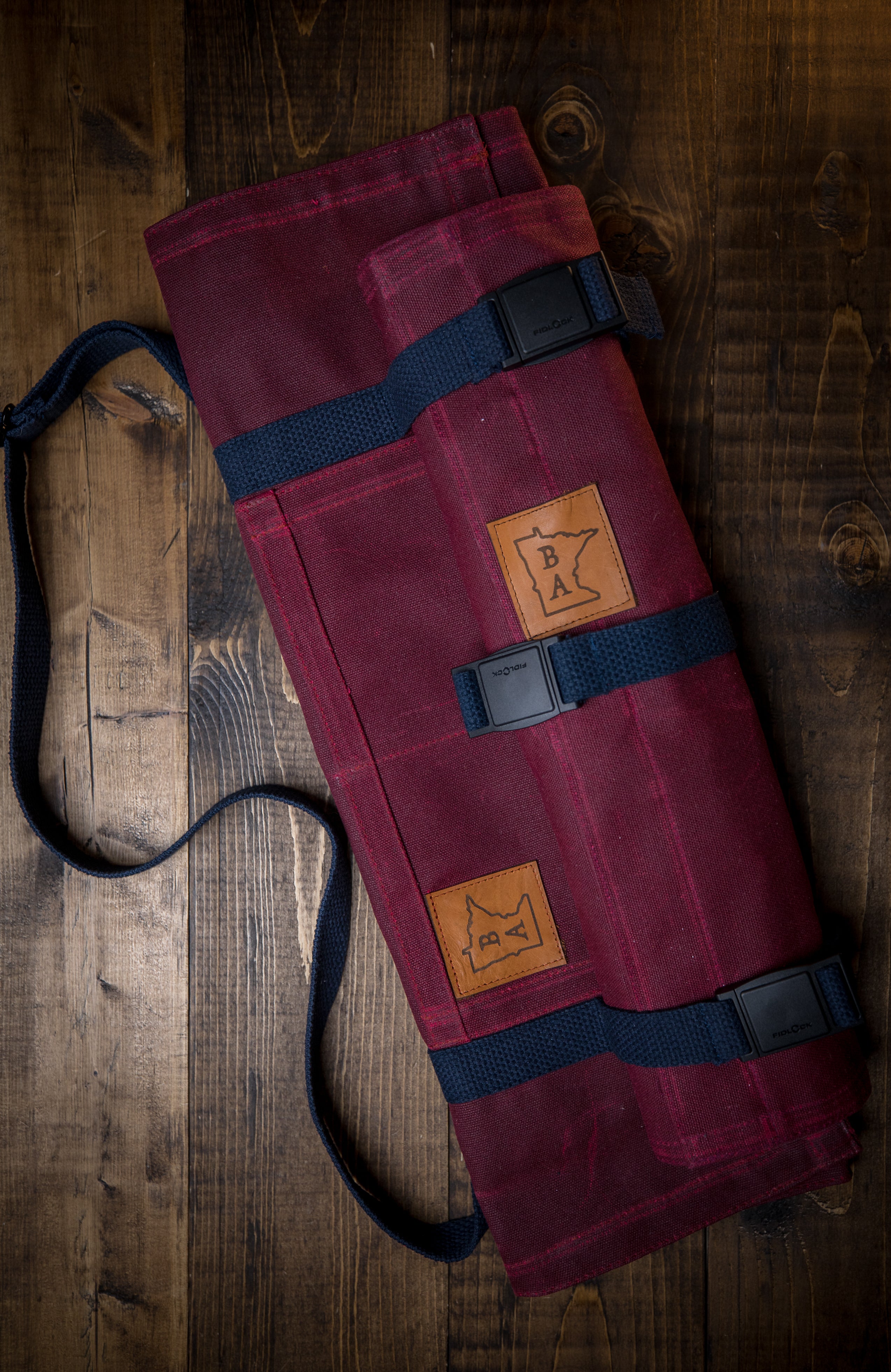 Main Squeeze burgundy cotton knife roll with a tex-wax outer shell canvas displayed on wooden surface. The interior of the knife roll is 50-50 Kevlar/Nomex blend for durability and protection. Shown with the matching set of the Side Hustle knife roll. Both designed by Craftmade Aprons, Minnesota.