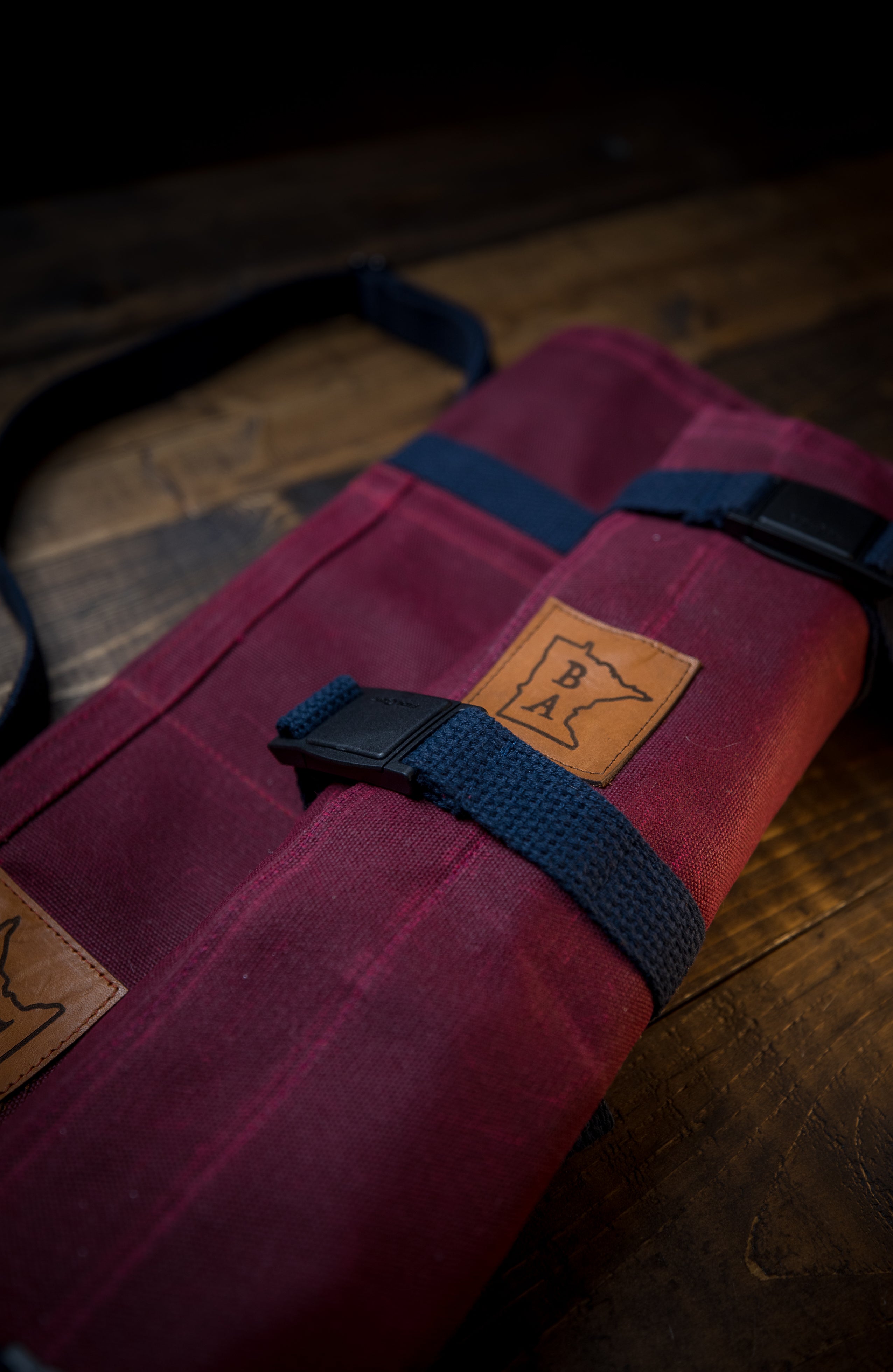     Product shot of the burgundy Side Hustle roll on a wooden surface. The cotton water repellent and flame resistant knife roll was designed by Craftmade Aprons, Minnesota.
