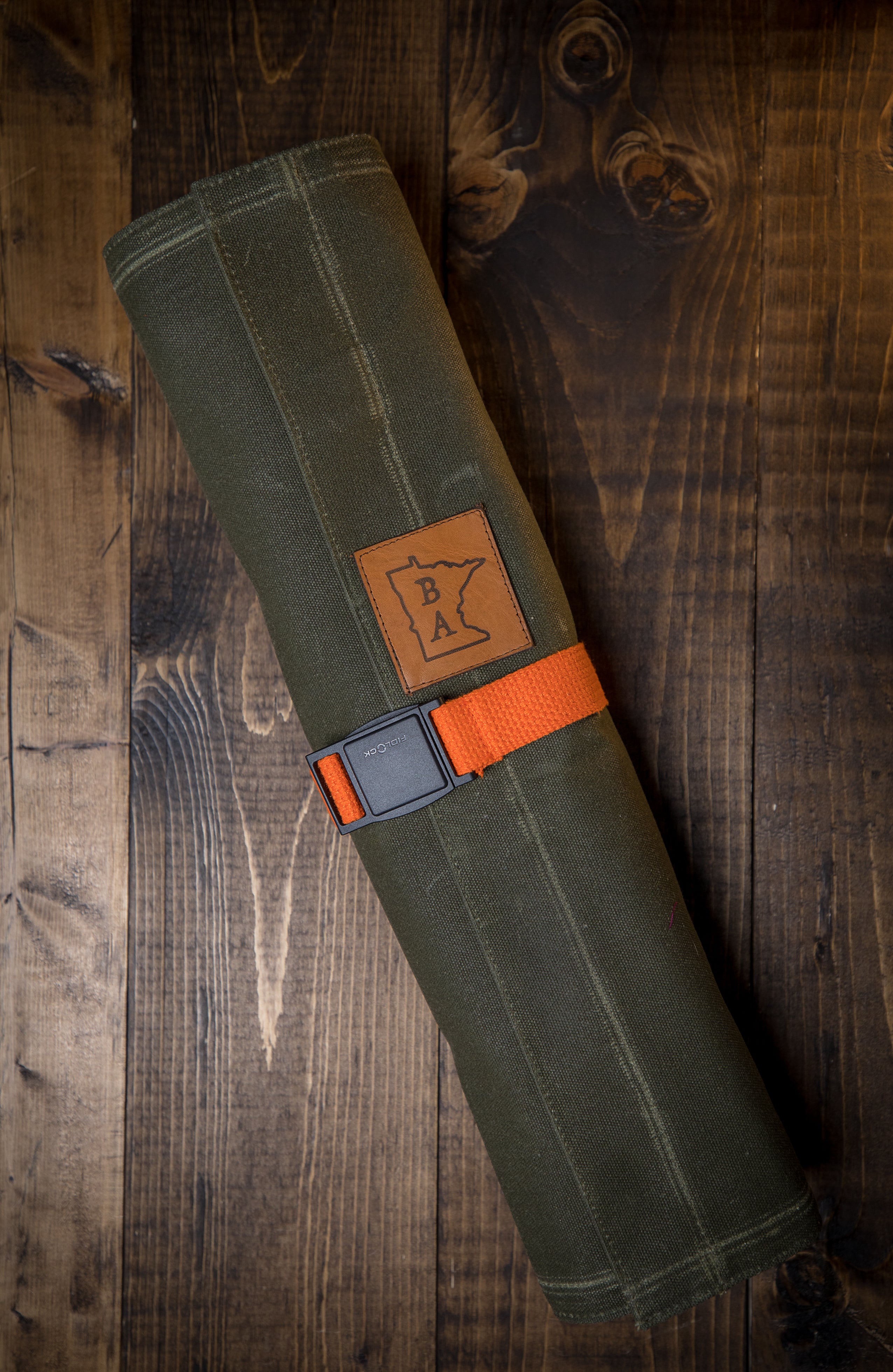   Cotton olive green knife roll protected in tex-wax folded with the orange strapping on a wooden surface. The knife roll design Side Hustle was manufactured in Minnesota by Craftmade Aprons
