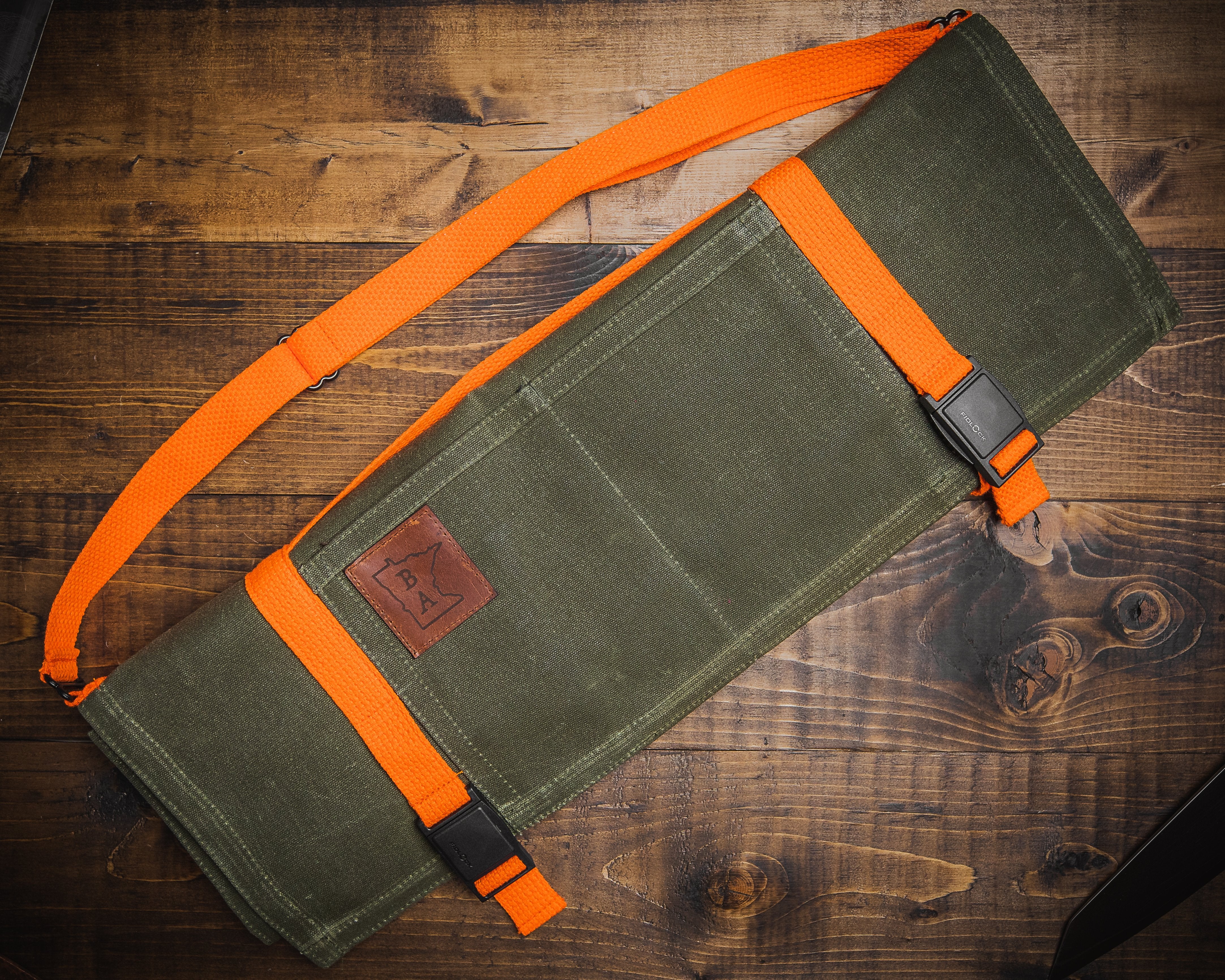 Olive green cotton knife roll Main Squeeze designed by Craftmade Aprons, Minnesota, laid on a wooden surface near a knife. 