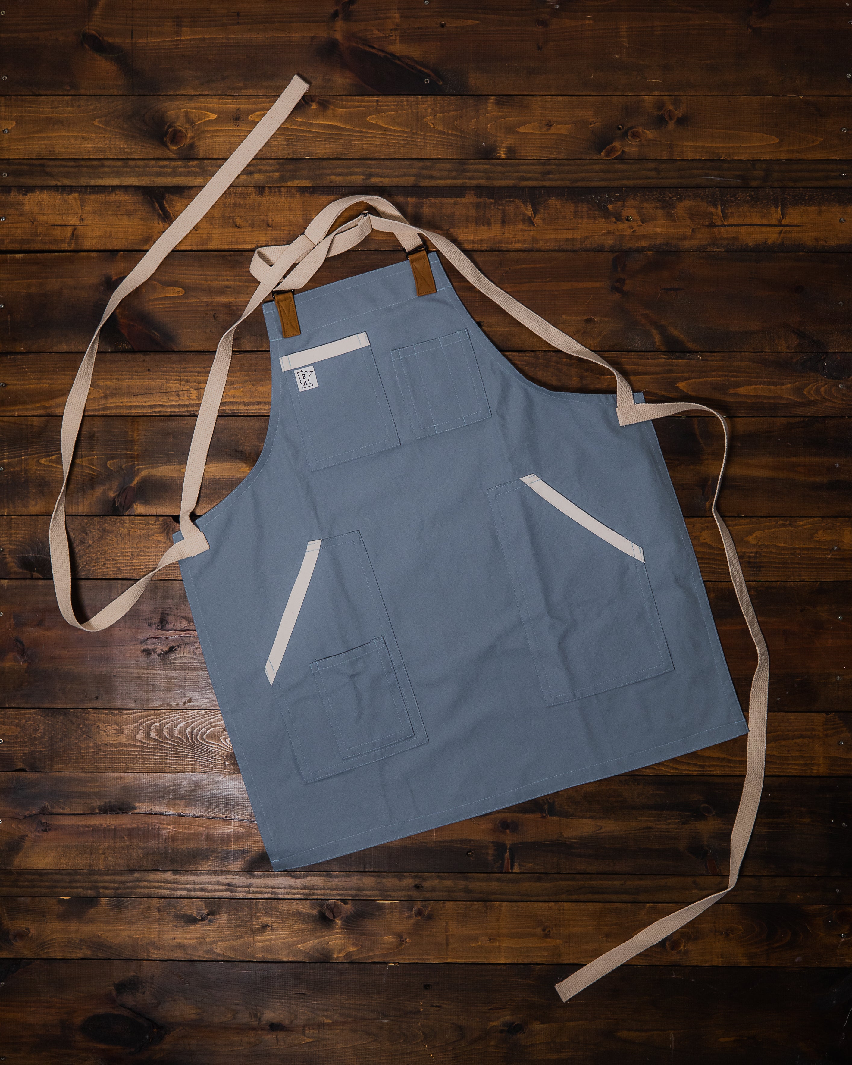 Cotton apron designed from 10oz duck cloth and has a muted blue base, cream accents on the pockets, and cream colored strapping laid out flat on a wooden surface. The apron design Blue Opal was manufactured in Minnesota by Craftmade Aprons.